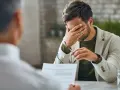 Mężczyzna z ręką na twarzy, siedzący przy biurku z dokumentami i okularami.