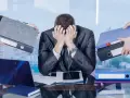 Pracownik w garniturze z rękami na twarzy, przytłoczony papierami i teczkami.